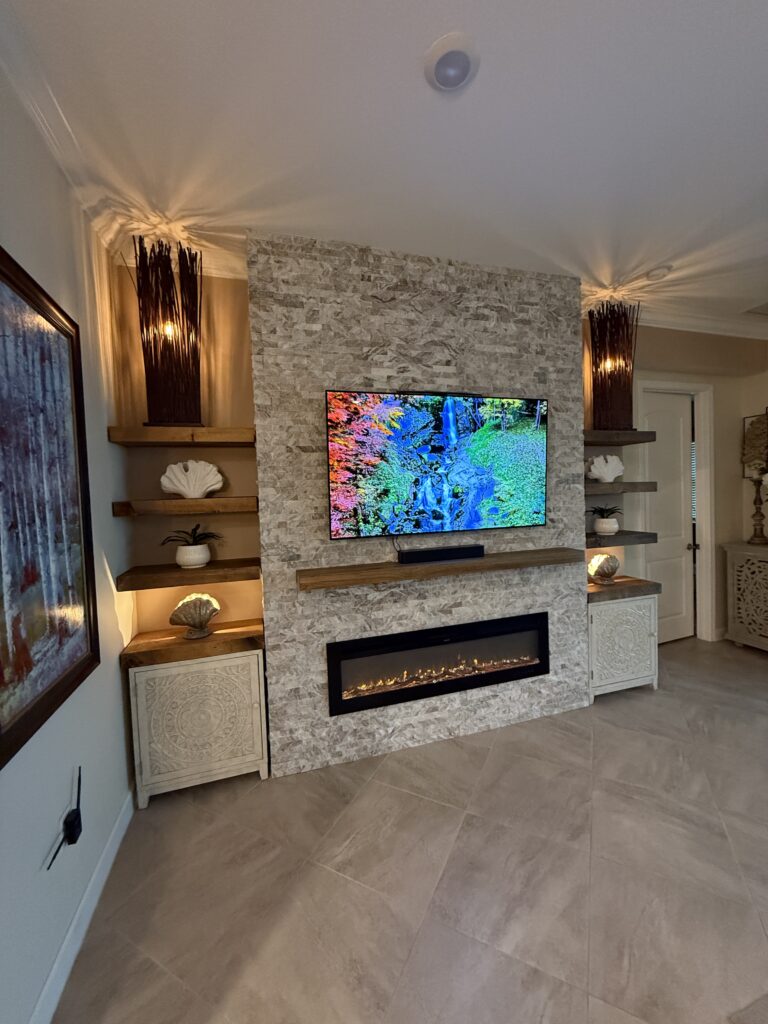 Angle view of stone fireplace TV wall with floating shelves, accent lighting, wood mantel, and electric fireplace