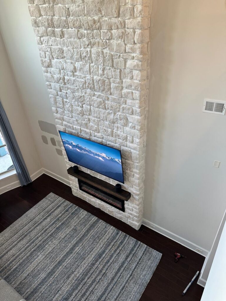 Angled view of floor to ceiling stone fireplace wall with TV, wood mantel, and electric fireplace