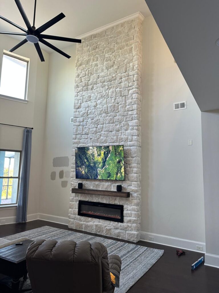 Floor to ceiling stone fireplace wall with TV, floating wood mantel, and linear electric fireplace in high ceiling living room