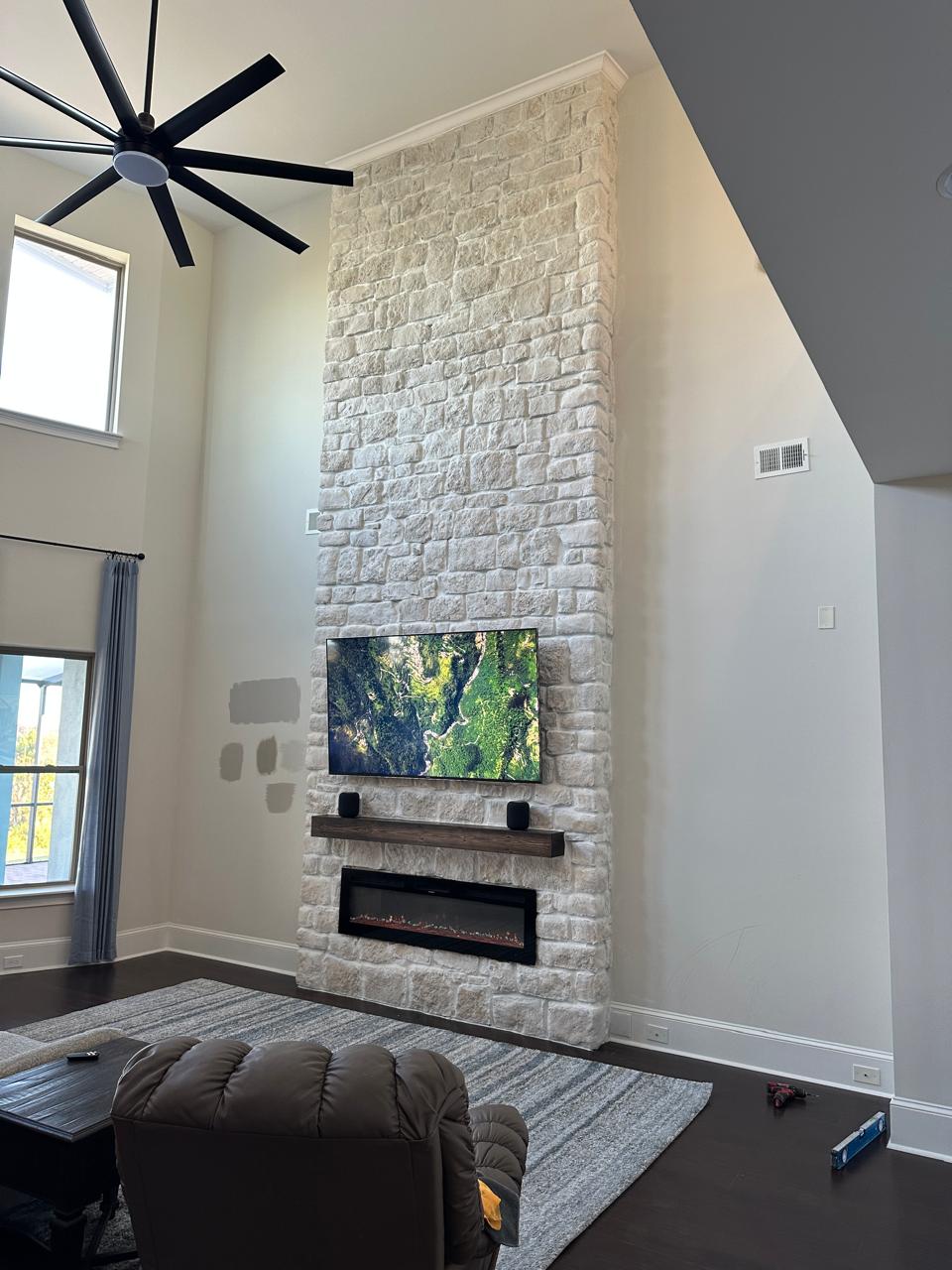 Floor to ceiling stone fireplace wall with TV, floating wood mantel, and linear electric fireplace in high ceiling living room