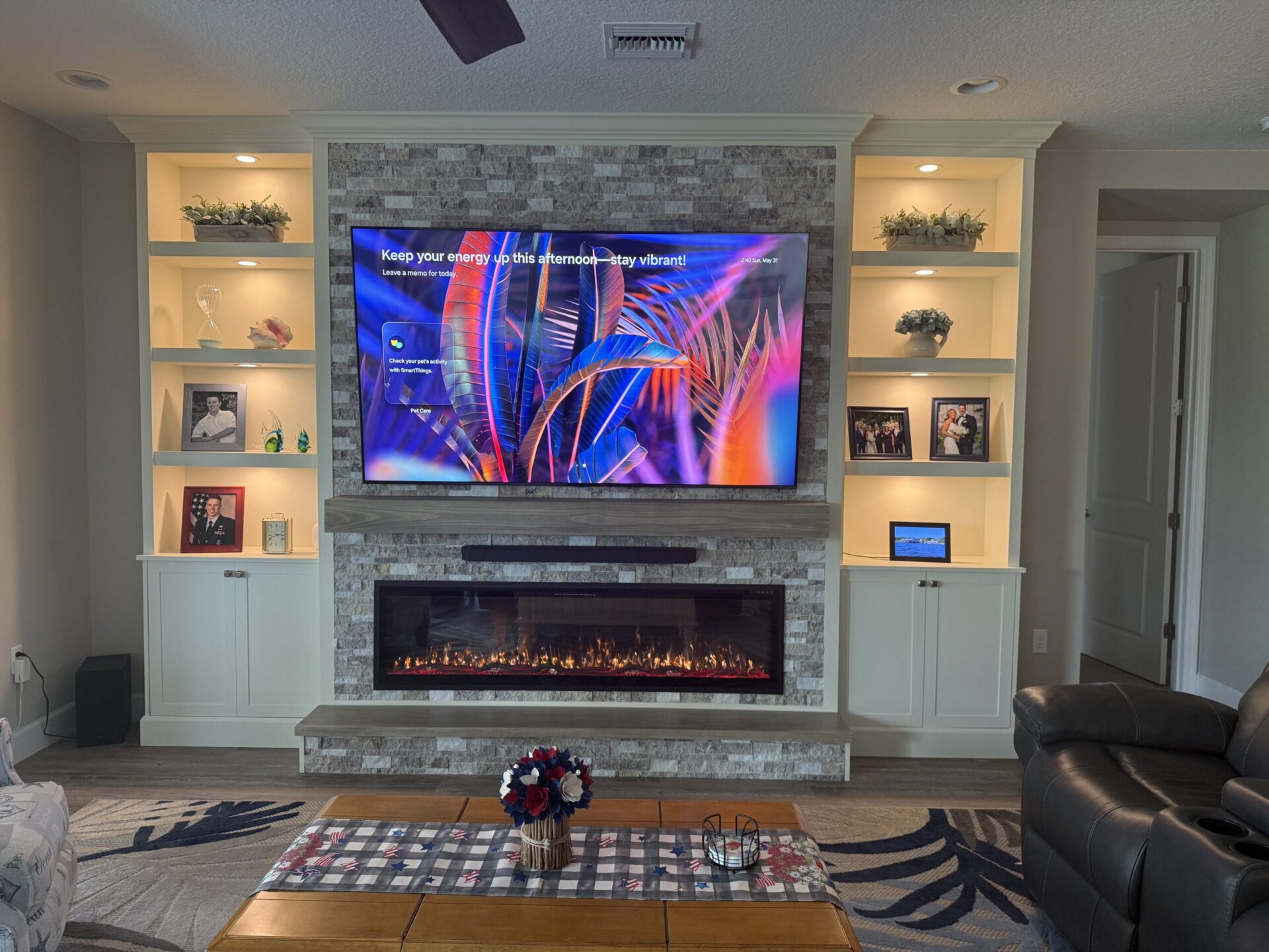 Custom media wall with integrated shelf lighting, stone fireplace surround, mounted TV, cabinets, and illuminated display areas